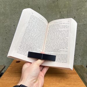 May include: A black book holder is holding open a paperback book. The book is open to a page with text about a spider and a group of creatures. The text reads, "The others laughed. "You were quite right, they'll soon put an end to that," hissed the angry spider climbing back onto the branch."