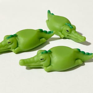 May include: Three green alligator figurines are displayed on a white surface. The alligators have a cartoonish design, with light green bodies, darker green spines, and closed eyes. The figurines are small and appear to be made of plastic.