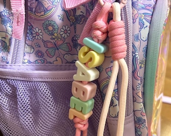 Puede incluir: Un colgante personalizado para mochila con cuentas cúbicas de colores pastel que forman el nombre ISABEL, sujeto a una mochila de color morado claro con un estampado de unicornios, mariposas y flores. Incluye un lazo de cordón rosa.