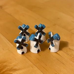 Peut inclure: Cinq moulins à vent miniatures en céramique blanche avec des pales peintes en bleu et noir. Les moulins à vent sont disposés en demi-cercle sur une surface en bois.