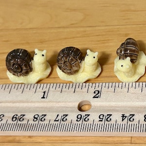 May include: Three miniature ceramic snail figurines with brown shells and white bodies. The snails are arranged in a row on a wooden ruler, with the ruler measuring 7.6 cm.