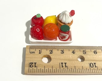 Puede incluir: Conjunto de comida en miniatura sobre un plato rectangular blanco. Incluye una manzana roja, fresa, limón, naranja, cupcake y un tarro de mermelada de fresa. Una regla que muestra centímetros está presente.