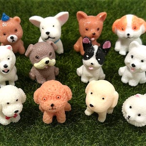 May include: A selection of miniature dog figurines in different breeds and colours. The figurines are in shades of brown, white, and black, with some featuring unique markings. They are displayed on a bed of green artificial grass.