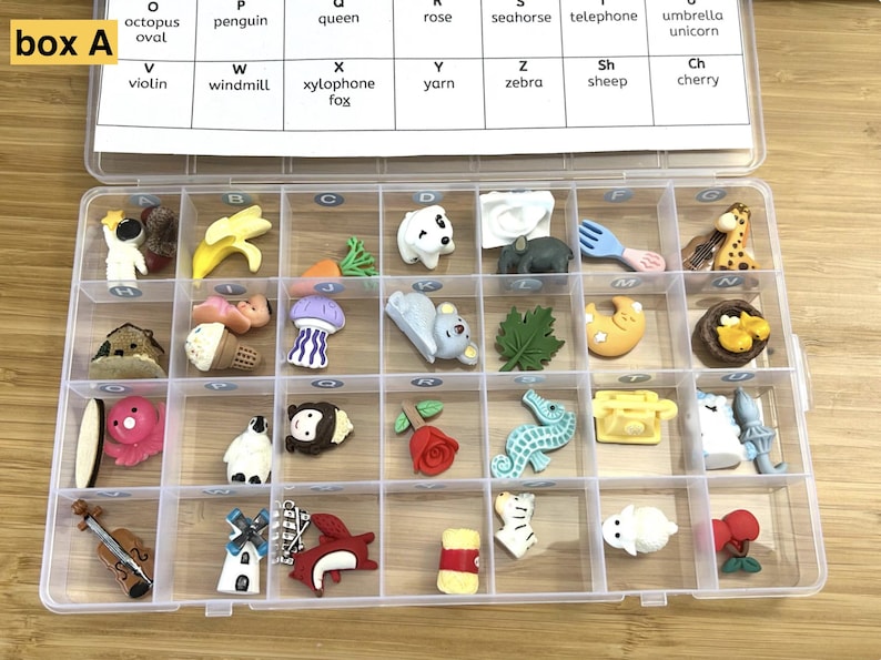Puede incluir: Una caja de almacenamiento de pl&aacute;stico transparente que contiene veinticuatro peque&ntilde;as figuras en miniatura de colores que representan diversos objetos y animales. Una tarjeta de papel blanco est&aacute; pegada a la tapa, con filas de letras y palabras correspondientes como octopus, penguin, queen, rose, seahorse, telephone, umbrella, unicorn, violin, windmill, xylophone, fox, yarn, zebra, sheep y cherry.