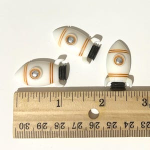 May include: Three white rocket ship-shaped embellishments with orange stripes and a silver-colored circle. The rockets have black bases and are arranged on a white surface next to a wooden ruler.