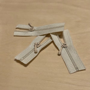 May include: Three off-white nylon zippers with matching plastic pull tabs. Each zipper features a woven fabric tape and a central coil mechanism. The pull tabs are teardrop-shaped and attached to a slider that moves along the teeth of the zipper.