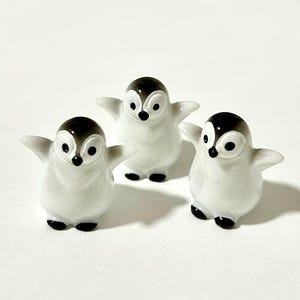 May include: Three small ceramic penguin figurines with black eyes and beaks. The penguins are white with black accents on their wings and backs. They are standing on a white surface.