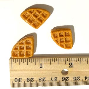 May include: Three miniature, golden-brown waffle pieces with a grid pattern are arranged on a white surface. A wooden ruler with inch markings is placed below the waffles, indicating their small size. The waffles appear to be food-themed miniatures.