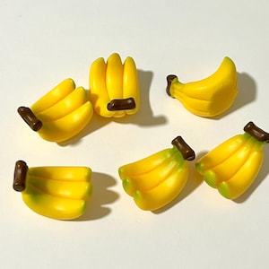 May include: Six miniature yellow banana bunches with brown stems. The bananas are arranged on a white surface.
