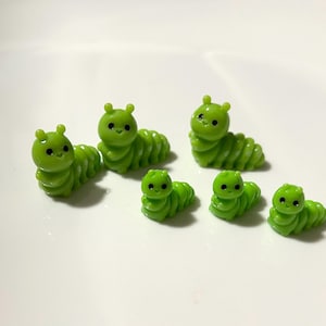 May include: Six small, green caterpillar figurines with black eyes and antennae. The caterpillars are in various poses, some upright and others crawling. The figurines are made of a glossy material and are set against a white background.