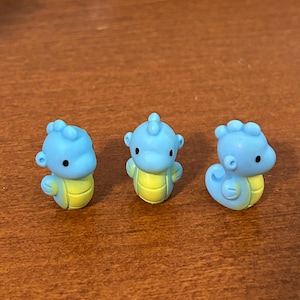 May include: Three small, light blue seahorse figurines with yellow bellies and black eyes are arranged on a brown surface. The seahorses have a cartoonish design, with rounded features and a playful appearance.