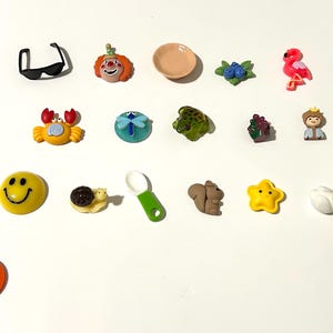 May include: A selection of small, decorative embellishments in various shapes and colours. The collection includes a smiley face, a crab, a flamingo, a strawberry, and the number 3. These miniature pieces are likely used for crafting or adding detail to projects.