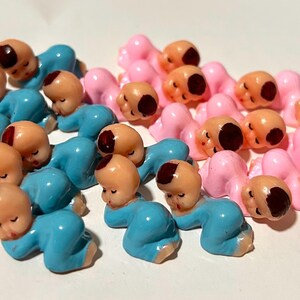 May include: A collection of small plastic figurines shaped like sleeping babies. The group consists of several figures in light blue and several figures in light pink. Each figurine features a peach-colored face with a painted brown hair patch.