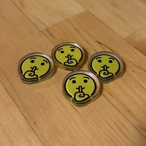 May include: Four yellow smiley face stickers with a finger over their mouths, symbolizing silence. The stickers are on a light brown wooden surface.