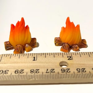 Peut inclure: Deux figurines miniatures de feu de camp. Chacune a une flamme aux tons orange et jaune, sur des bûches en bois marron. Les figurines sont placées sur une surface blanche à côté d'une règle. La règle montre des mesures en centimètres.
