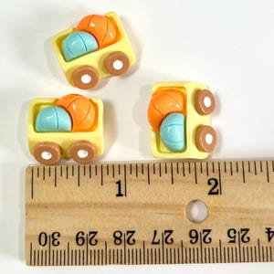 May include: Three yellow toy truck charms, each carrying a blue and orange ball. The charms have brown wheels and are arranged on a white surface. A wooden ruler with inch markings is in the foreground.