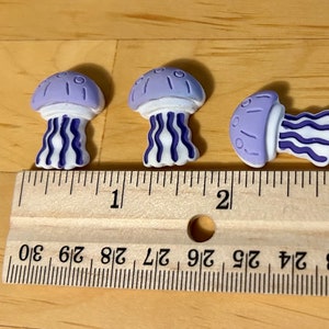 May include: Three purple and white jellyfish charms, each approximately 2.5 cm in size, are arranged on a wooden surface next to a ruler.