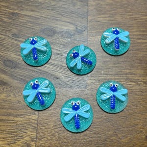 May include: Six decorative dragonfly embellishments. Each dragonfly has light blue wings and a dark blue body, set on a teal green circular base. The embellishments are arranged on a wooden surface.