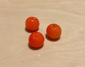 Puede incluir: Tres pequeñas naranjas artificiales redondas, texturadas y de color naranja sobre una superficie de madera marrón claro.