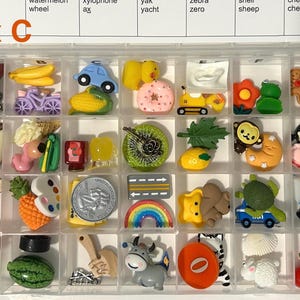 Puede incluir: Un contenedor de almacenamiento de pl&aacute;stico transparente con veintiocho compartimentos llenos de varios dijes de resina en miniatura, incluyendo frutas, animales, veh&iacute;culos y alimentos. La tapa presenta una cuadr&iacute;cula impresa con el texto box C.