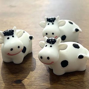 May include: Three small, white cow figurines with black spots and black hair. The cows have smiling red mouths and are arranged on a wooden surface. These are whimsical decorative items.