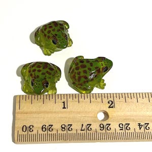 May include: Three small, translucent green frog figurines with brown spots. The frogs are positioned on a white surface next to a wooden ruler. The ruler shows measurements in inches.