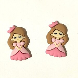 May include: Two princess-shaped embellishments. Each princess has brown hair, a pink crown, and a pink dress. The princess is smiling with her hands clasped in front of her. These embellishments are suitable for crafts.