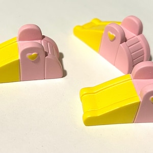 May include: Three miniature playground slides featuring a bright yellow sliding surface and a pastel pink frame. Each slide includes a small yellow heart detail on the side panel and a set of moulded steps leading to the top of the slide structure.