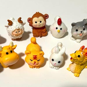 Peut inclure: Ensemble de douze figurines colorées représentant les animaux du zodiaque chinois. Chaque figurine est unique, avec un dragon rouge, un mouton blanc et une souris bleue, entre autres.