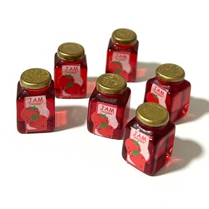 Peut inclure: Sept petits pots carrés de confiture de fraises avec des couvercles dorés. Chaque pot porte une étiquette avec le mot "JAM" et une image de fraises. La confiture est d'un rouge profond.