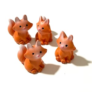Peut inclure: Quatre petites figurines de renard en céramique orange avec des accents blancs. Les renards sont assis sur une surface blanche.