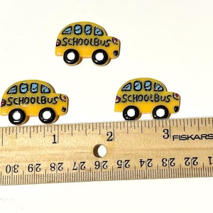 Puede incluir: Tres adornos amarillos con forma de autobús escolar con ruedas negras y la palabra "SCHOOLBUS" impresa. Una regla de madera se coloca junto a los adornos. Los adornos miden aproximadamente 2,5 cm de largo.