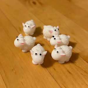 May include: Six white ceramic sheep figurines with black eyes and pink noses. The sheep are arranged in a circle on a wooden surface.