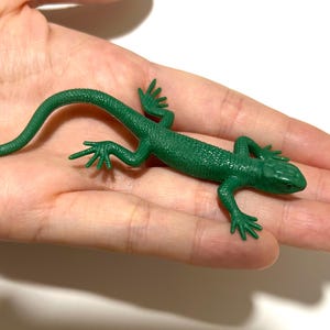 Peut inclure: Une figurine de lézard verte avec une surface texturée. Le lézard est posé sur une paume, mettant en valeur ses détails, dont ses pattes, sa queue et sa tête. La figurine est de couleur verte vive.
