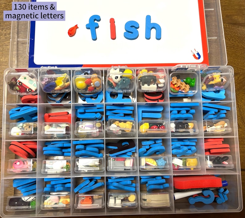 Puede incluir: Un contenedor de almacenamiento de pl&aacute;stico transparente con m&uacute;ltiples compartimentos llenos de peque&ntilde;os objetos coloridos y letras magn&eacute;ticas. La tapa blanca muestra el texto 130 items & magnetic letters y la palabra fish.