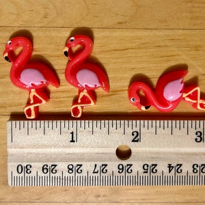 May include: Three pink plastic flamingo figurines, each about 2.5 cm tall, are lined up on a wooden ruler. The ruler is marked in inches and shows the figurines are about 2.5 cm long.