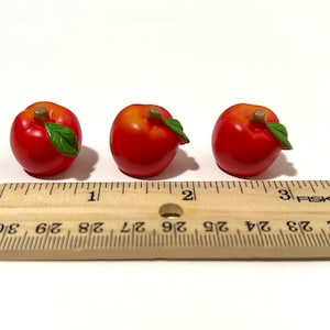 May include: Three miniature red apples with green leaves and brown stems are arranged in a row. A wooden ruler with centimetre markings is placed below the apples, showing measurements up to 10 cm.