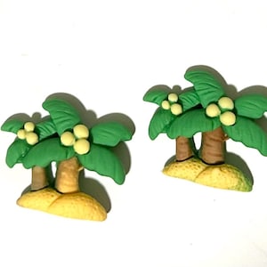 May include: Two miniature palm tree figurines. Each tree has a brown trunk, green fronds, and yellow coconuts. They are set on a yellow base, resembling a small island. The background is white.