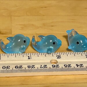May include: Three translucent blue dolphin figurines with black eyes are arranged in a row. A wooden ruler with centimetre markings is placed in front of the dolphins. The dolphins are small, decorative items.