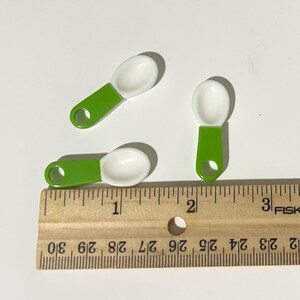Peut inclure: Trois petites cuillères à mesurer avec des cuillères blanches et des manches vert vif. Les manches ont un trou pour les suspendre. Les cuillères mesurent environ 2,5 cm de long. Une règle en bois est au premier plan.