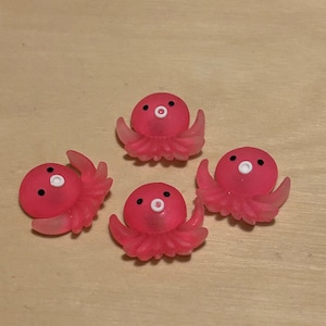 May include: Four pink octopus-shaped figurines with white eyes and a small white circle in the centre of each octopus. The octopus figurines are on a light brown surface.