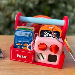 May include: A red wooden toy toolbox with a light blue handle, containing flashcards and a pink toy radio. The toolbox has the name "Parker" on it. A stack of flashcards is on the table next to the toolbox. The flashcards have the text "I'M" on them.