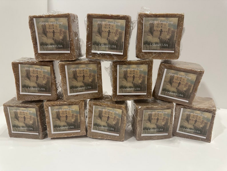 Puede incluir: Una pila de 12 barras marrones de jab&oacute;n de laurel de Alepo, cada una envuelta en pl&aacute;stico y etiquetada con el texto "Aleppo Laurel Soap" y "LABASH USA".
