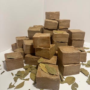 May include: A collection of square, light brown soaps, some with dried bay leaves on top, arranged on a white surface. The soaps are stacked in a pile, with additional leaves scattered around the base, creating a natural, earthy aesthetic.