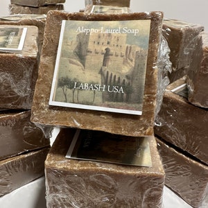 Puede incluir: Una pila de barras de jab&oacute;n de laurel de Alepo de color marr&oacute;n envueltas en pl&aacute;stico. La barra superior tiene una etiqueta que dice "Aleppo Laurel Soap" y "Labash USA" con una ilustraci&oacute;n de un edificio y &aacute;rboles.