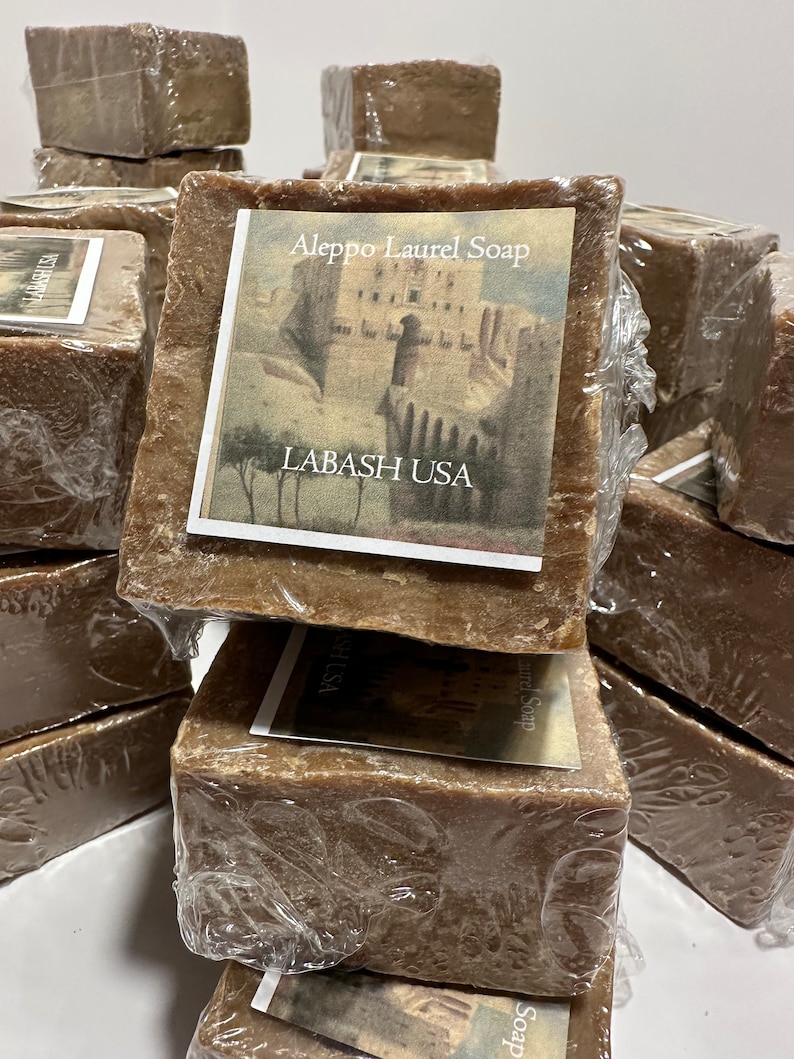 Puede incluir: Una pila de barras de jab&oacute;n de laurel de Alepo de color marr&oacute;n envueltas en pl&aacute;stico. La barra superior tiene una etiqueta que dice "Aleppo Laurel Soap" y "Labash USA" con una ilustraci&oacute;n de un edificio y &aacute;rboles.