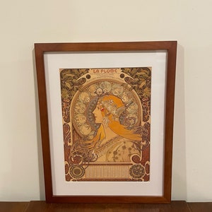 May include: A framed print of a vintage poster for "La Plume", a French magazine. The poster features an Art Nouveau design with a woman's profile, intricate floral patterns, and a circular design with zodiac symbols.