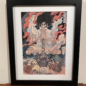 May include: Framed artwork featuring a detailed illustration of a seated figure with a fierce expression, surrounded by flames and mythical creatures. The artwork is in a black frame with a white mat.