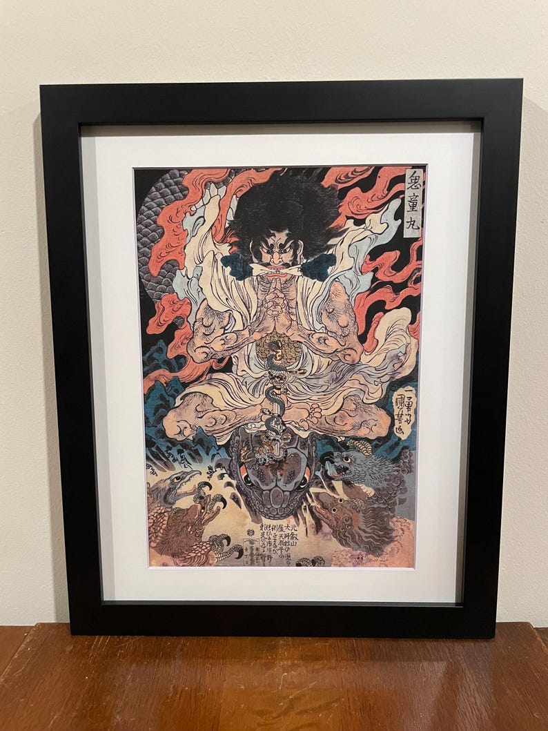 May include: Framed artwork featuring a detailed illustration of a seated figure with a fierce expression, surrounded by flames and mythical creatures. The artwork is in a black frame with a white mat.