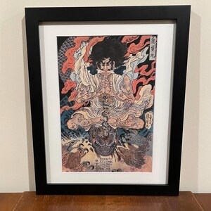 May include: Framed Japanese artwork featuring a detailed illustration of a deity with a fierce expression, surrounded by flames and mythical creatures. The artwork is in a black frame with a white mat, showcasing the vibrant colors and intricate details of the print.